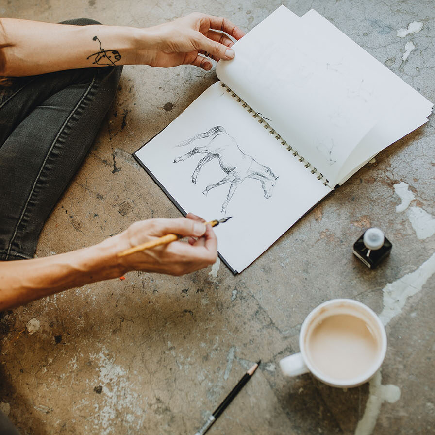 sketchbook on concrete floor with a horse in pen and ink and coffee cup next to it